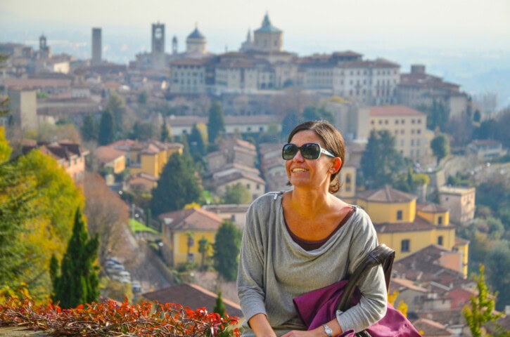 A woman wearing sunglasses and a gray top sits outdoors with a European town in the background on a sunny day in Italy. She holds a purple bag and smiles slightly.