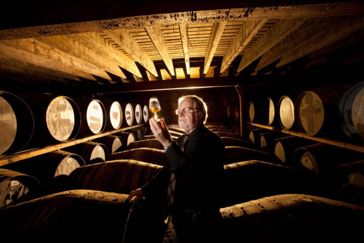 A person stands in a dimly lit cellar in Scotland, surrounded by barrels, holding a glass of liquid up to the light for examination. This hidden gem invites you to discover the rich heritage of Scottish craftsmanship—an unforgettable experience for any travel enthusiast.