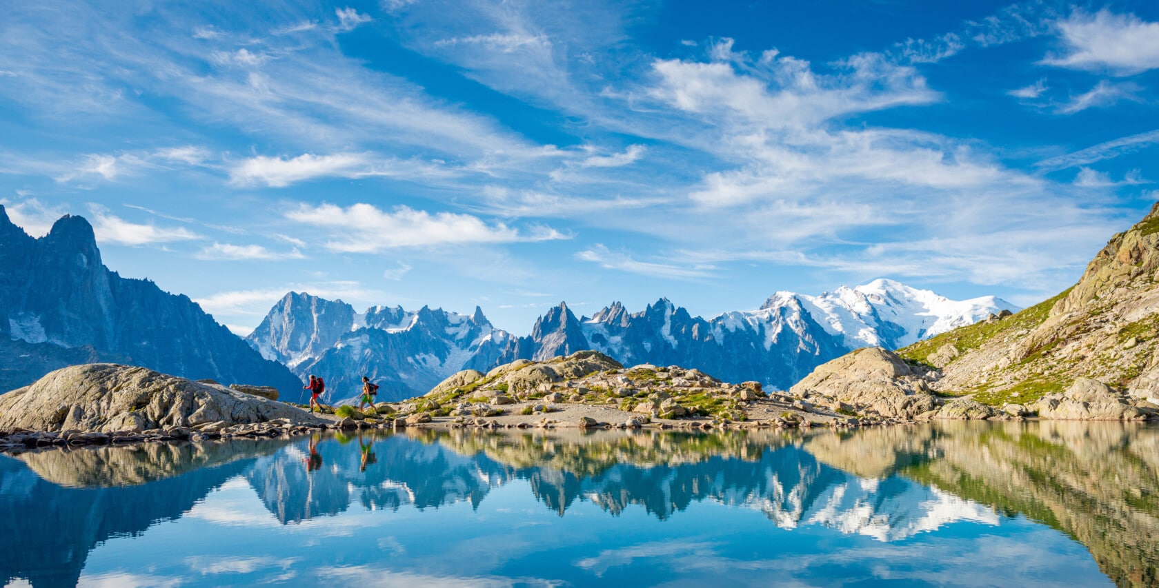 Two hikers walk on a rocky path at the bottom of a clear lake reflecting snow-capped mountains and a bright blue sky with scattered clouds, creating scenes worthy of an awards archive.