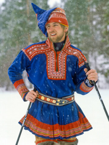A person in traditional Sámi clothing stands outdoors in snowy surroundings, holding ski poles, typical of Northern Europe's Scandinavian countries.